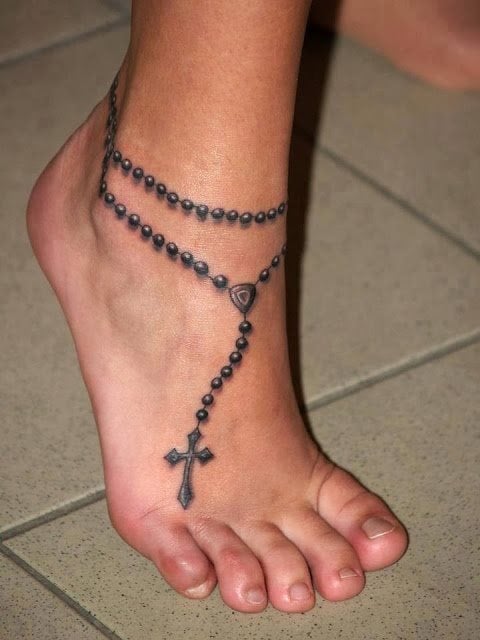 Clsico diseo de un rosario enlazado al tobillo de esta joven y con la cruz reposando sobre el empeine del pie de la misma con unos brillos en las bolas que le dan cuerpo al diseo