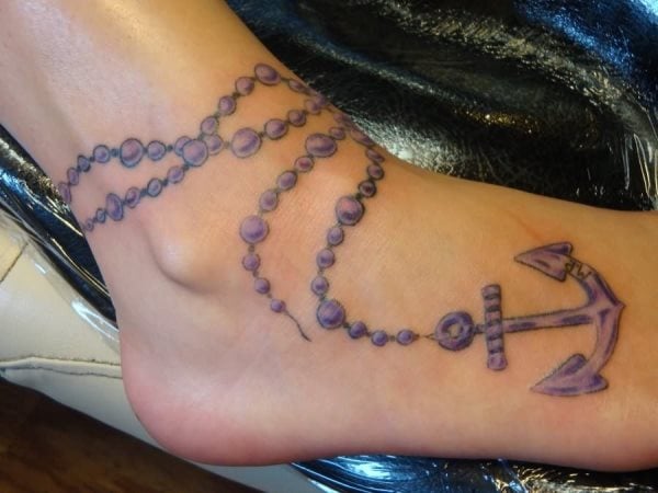 Tattoo de un rosario que rodea el tobillo y cae sobre el empeine y termina el ancla