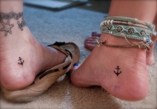 Sencillo tatuaje de un ancla en tonos negros en la parte del taln del pie