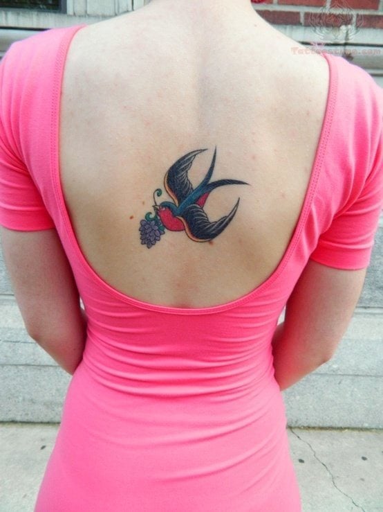 En el centro de la espalda, tatuaje de una golondrina de estilo old school que lleva un ramillete de flores en su pico