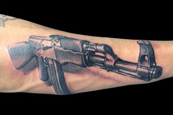 Tatuaje en el antebrazo de una metralleta de gran tamaño, en la que podemos apreciar todos los detalles de esta arma, desde el cañón, la empuñadura, el gatillo, el cargador y un largo etcétera de accesorios que no se han dejado sin tatuar