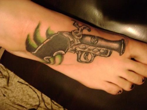 Tatuaje de una pistola clásica sobre el pie y en el que se ha dibujado unos trazos negros y verdes bajo la culata de la pistola