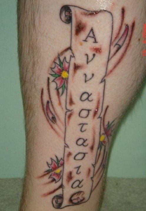 Tatuaje en la pierna de un lazo en forma de papiro al que se le ha inscrito una palabra dentro y que se ha adornado con unas flores de colores de pequeño tamaño y algunas cenefas y sombreados bajo el lazo