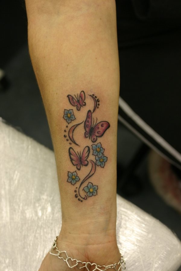 Este tatuaje ubicado en el antebrazo de esta chica tiene como motivo un conjunto de flores celestes y amarillas y mariposas rosas en lo que parece ser una brisa de aire
