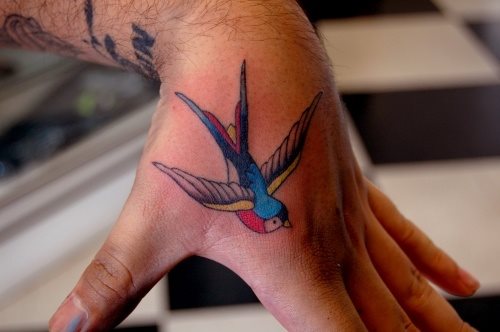 Tonos azules, amarillos y rojizos utilizados para hacer este tatuaje de una golondrina en la mano
