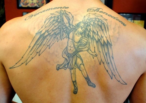Tatuaje de un ángel guerrero sobre la espalda con grandes alas, con un elegante plumaje y al que se le ha realizado una frase en la parte superior del tatuaje, nos gusta especialmente el fondo tan difuminado que se le ha trazado bajo el ángel