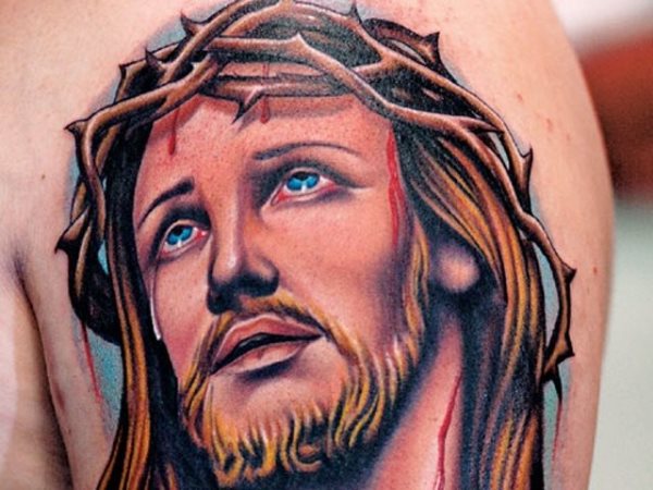 Tatuaje a color de un Cristo con el pelo rubio y ojos azules, del que de la corona de espina se desprenden gotas de sangre y al que se le ha tatuado unas lágrimas en uno de sus ojos, un Cristo diferente al que estamos acostumbrados a ver a lo largo de la historia