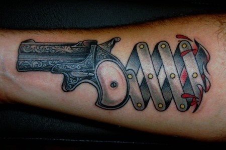 Peculiar tatauje de pistola sobre el brazo, en el que destacan las filigranas del gravado de la pistola y el elástico de metal que parece salir de la propia carna del tatuado, para ello se han dibujado unas cuantas gotas de sangre