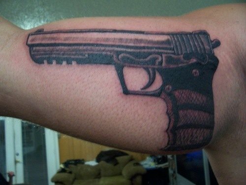 Tatuaje de una pistola de gran calibre en el interior del bíceps, es un tatuaje realizado a color negro y en la que los acabado de la empuñadora formando una rejilla, para nuestro gusto, no está demasiado bien conseguidos, aunque el tatuaje en sí no es malo del todo