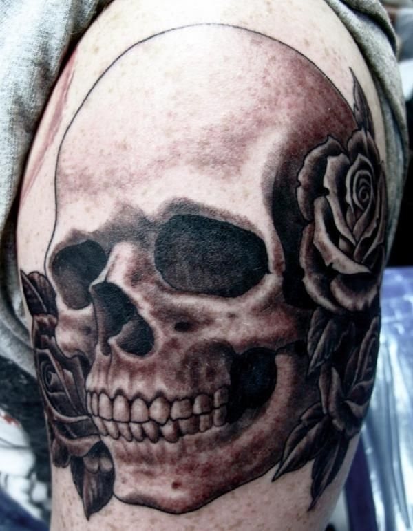 Tatuaje de calavera y rosas negras ocupan todo el brazo de este hombre, gracias al colorear las cavidades de los ojos en negro se ha conseguido alcanzar una buena profundidad de los ojos y los pétalos de las rosas también están muy bien conseguidos