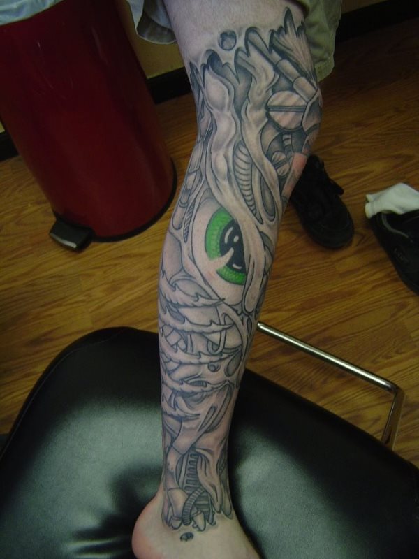Tatuaje en la pierna de lo que parece una especie de robot dragón con tornillos y cables, al que se sólo se le ha tatuado en color un gran ojo verde y negro, con buenos brillos conseguidos en la parte negral del ojo