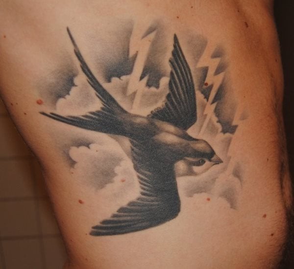 Tatuaje de una golondrina en pleno vuelo con las alas bien abiertas y bien definidas, que se ha completado con un fondo en color negro de nubes y truenos, un buen resultado en una zona muy acertada y la verdad es que es un tattoo de gran tamaño