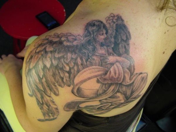 Tatuaje de una ángel de gran belleza con unas plumas preciosas que forman las alas y una túnica que cubre todo su cuerpo que ha sido elaborada con gran perfección en el juego de las curvas y sombreados