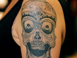 Calavera sobre el brazo con un aspecto aparentemente simpático y para el que se han utilizado grandes ojos y un relleno de dibujos en la parte ósea