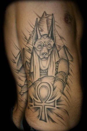 Otro diseño de Anubis rodeados de otros símbolos egipcios, en el que se destaca la cruz típica egipcia y las pirámides egipcias al fondo, dotando al tatuaje de una gran profundidad y simbolismo