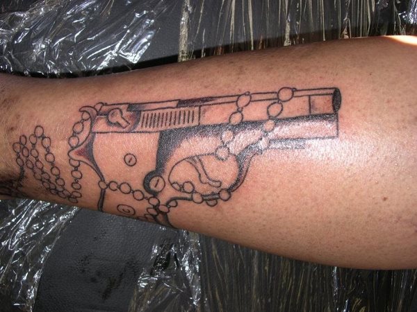 Tatuaje de una pistola rodeada por un rosario, un tatuaje un tanto contradictorio, pero muy utilizado en muchas zonas de América, ya que son muchos pistoleros los que buscan la protección cristiana para que a ellos no les pase lo mismo que ellos hacen