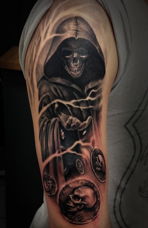 Tatuaje de la representación de la muerte tatuado sobre el brazo, en color negro, como no podría ser de otra manera y bajo esta representación se encuentran unas monedas que parecen estar volando y que se han acuñado con una calavera lateral, emulando a que es la propia muerte quien está tirando al aire las monedas