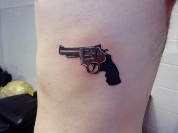 Pequeño diseño de un arma de tipo revolver a color negro, tatuado sobre el costado de este hombre