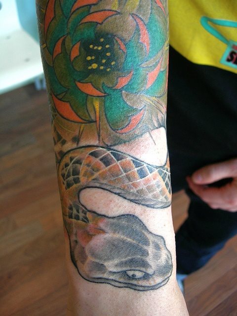 Tatuaje muy colorido de una serpiente y una flor, a pesar de que la serpiente y el relleno de las escamas tienen unos colores muy acertados, nos encanta la flor y la profundidad que se ha conseguido, gracias a la forma que hacen las hojas y al sombreado que se le ha hecho en la parte del estigma