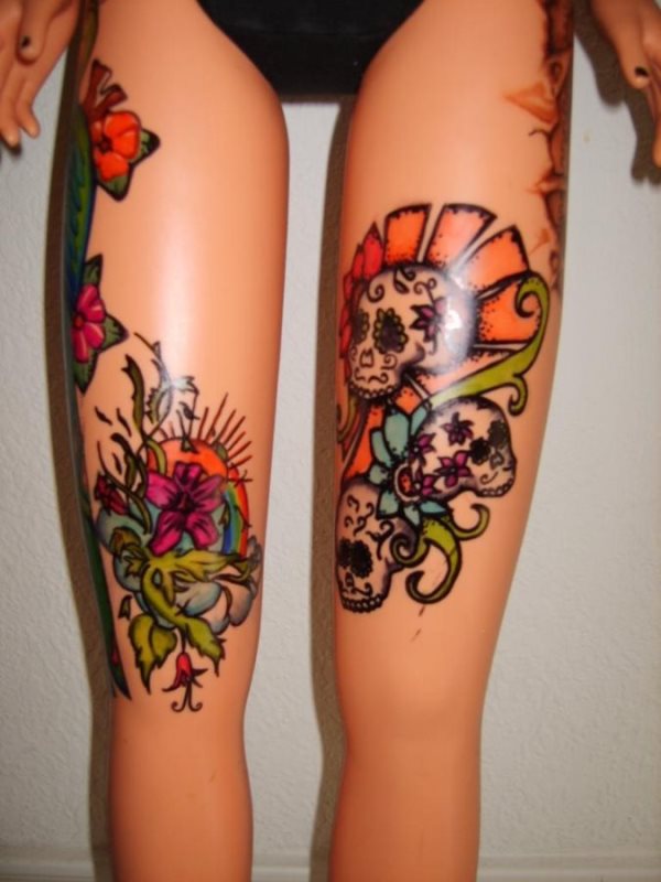 Tatuaje de calaveras de estilo mexicano, bordeadas con gran colorido de trazos naranjas y cenefas verdes, que emulan a las ramas de algunas flores, estamos ante un tatauje muy bonito que como podemos ver no es real, al igual que la muñeca sobre la que se han realizado los tattoos