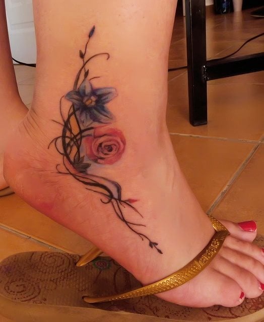 Tatuaje en el tobillo de unas flores pequeñas, acompañadas por unos ramilletes de líneas que se van cruzando, un trabajo muy bonito sobre el tobillo y del que nos gusta mucho a calidad del dibujo de la flor rosa