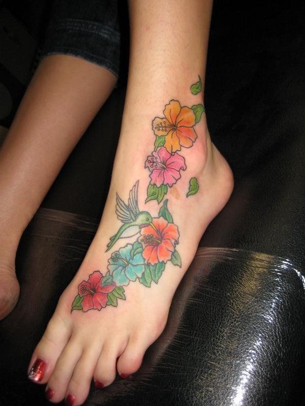 Tatuaje de flores que baja desde el tobillo hasta el pie y donde podemos observar como se ha tatuado un pequeño pajarillo de color turquesa que tiene su pico sobre los estambres, un divertido y elegante tattoo muy colorido