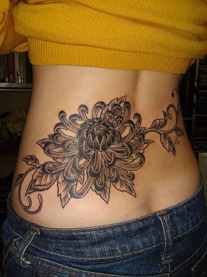 Tatuaje en la cintura de una mujer, en la que se puede apreciar una enorme flor a color negro, en el que los pétalos van haciendo una maravillosa forma y está acompañada de unas hojas, en el que las ramas hacen las veces de las cenefas del tattoo
