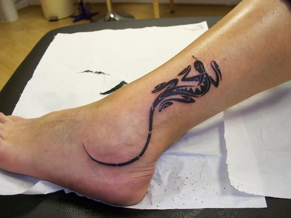 Tobillo tatuado por un pequeño lagarto que tiene una cola muy larga qeu rodea el tobillo