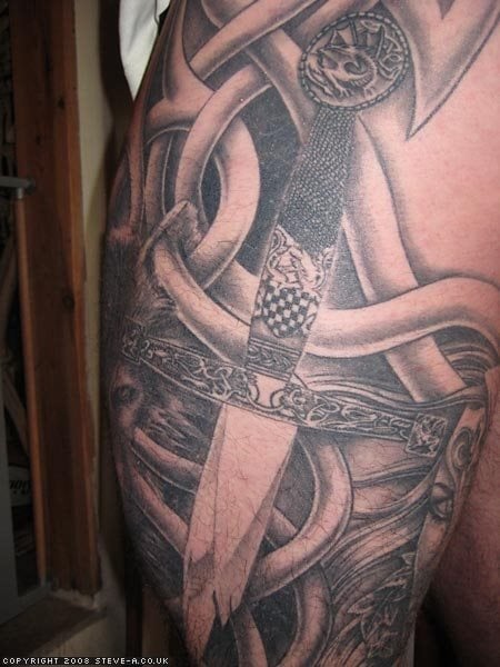 Tatuaje de una pequeña daga con motivos de dragones y gran tallado tanto en la hoja como en la empuñadura, tatuado sobre un enorme tribal con un espectacular fondo negro que favorece a los trazos conseguidos con motivos tribales