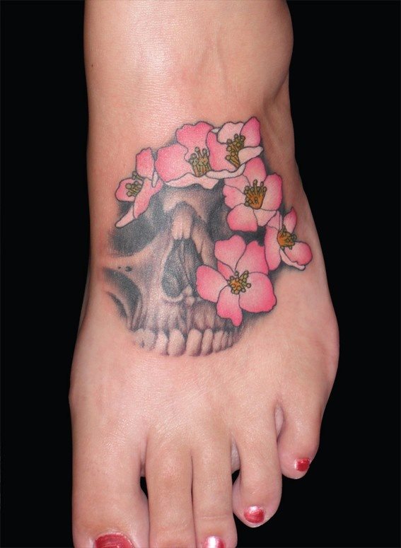 Tatuaje en el pie de una calavera rodeada por unas cuantas de flores de pétalos color naranja, que casi envuelven a la calavera impidiendo que se vea en su totalidad, un tatuaje muy bonito para la zona escogida