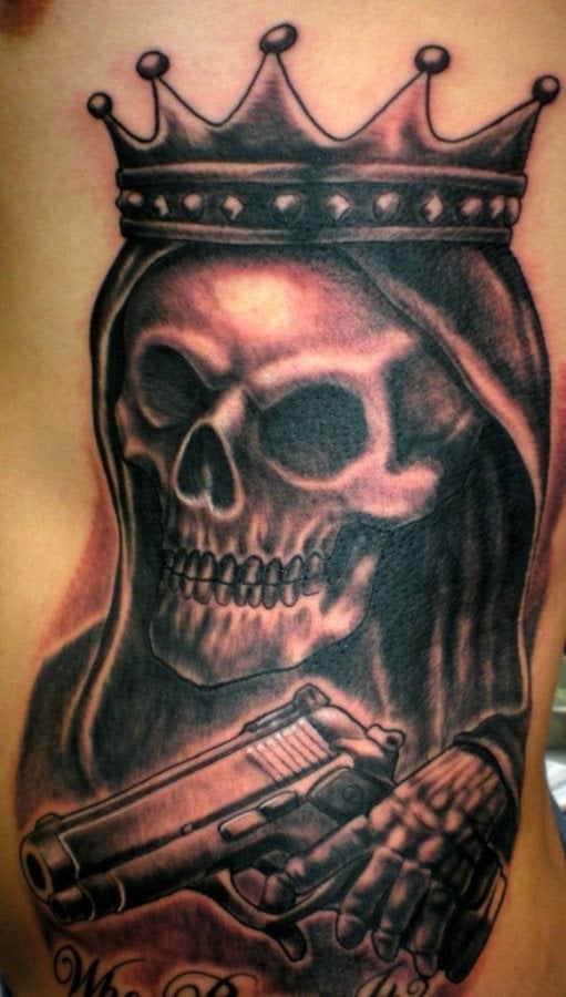 Tatuaje de una calavera con capucha y corona de cinco puntas que porta en sus manos una pistola de gran calibre, un tatuaje sin duda aterrador y que gustará a casi todos los amantes de los tatuajes de calaveras