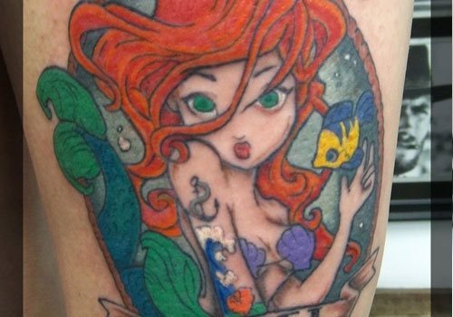 Diseño infantil de La Sirenita sobre el brazo, la sirenita tiene unos grandes ojos de color verde y boquita de piñón mandando un beso a todos aquellos que se fijen en este colorido tatuaje en el que los peces, el agua y las hojas están muy bien marcadas por la cuerda que rodea a todo el tatuaje, excelente el detalle de la conchas como parte de arriba del bikini de la sierinita tatuada