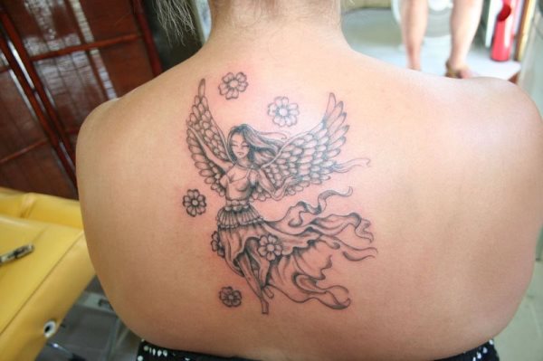 Tatuaje de un ángel caracterizado por una larga flore con marcados detalles, una camiseta de tirantas y unas alas con plumas pequeñas y anchas, de la que se ve sobresalir el largo pelo de este ángel, además al tatuaje se le han ido añadiendo unas pequeñas flores que le dan un acabado muy conseguido