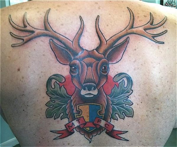 Tatuaje de un ciervo que parece el escudo de algún club de prestigio o una universidad prestigiosa, sin duda un resultado muy bonito y un logo que nos gusta bastante, sobre todo por el fondo del cielo naranja y el lazo con las iniciales