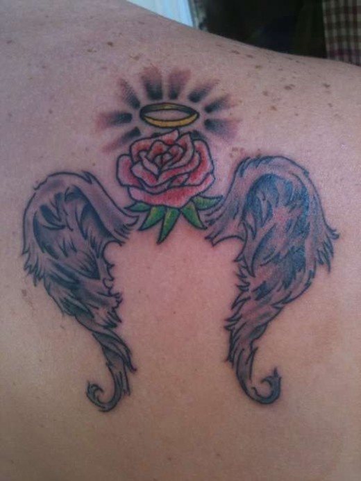 Tatuaje en el omóplato de unas alas un tanto poco definidas, para la que se ha trazado una rosa roja en el centro, con pequeñas hojas verdes y en la parte superior, una corona dorada de la que se desprenden algunos destellos, un tatuaje que con algún que otro repaso podría quedar algo mejor