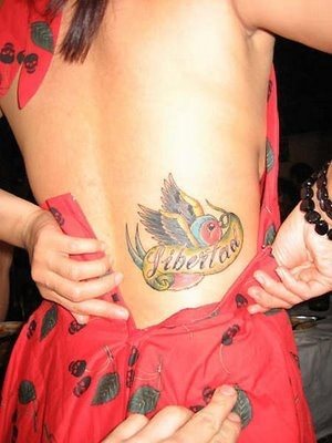 Tatuaje en la parte baja de la espaalda de una golondrina preciosa y muy colorida que sobre su pico lleva un lazo con la palabra 