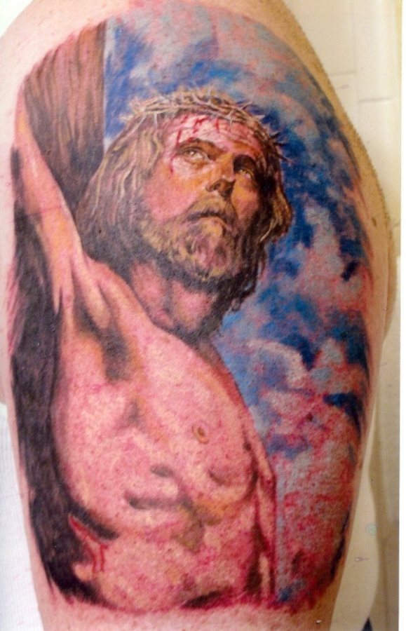 Tatuaje a todo color de un Cristo crucificado miurando hacia un lado, al que se le ha añadiedo un espectacular y buenísimo fondo de nubes blancas y cielo azul, sin duda estamos ante uno de los mejores trabajos de tatuajes cristianos que hemos visto