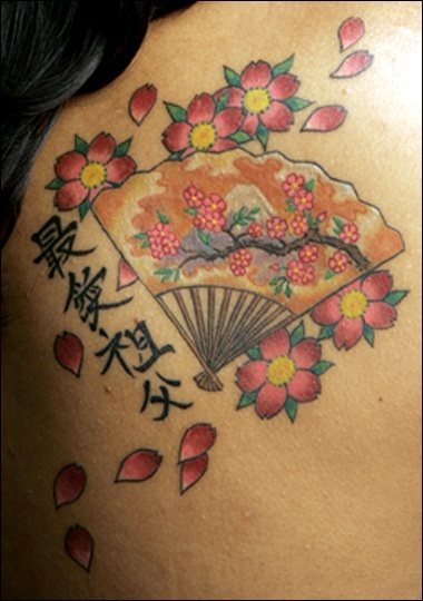 Tatuaje de un abanico de estilo oriental rodeado por un conjunto de flores a las que se le están cayendo algunos pétalos y que ha sido completado con varias letras chinas en un lateral del abanico