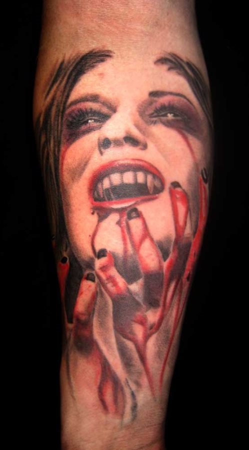 Tatuaje de una mujer vampiro con grandes colmillos que se toca la cara con sus manos manchadas de sangre, tal vez porque está disfrutando del momento de éxtasis tras haber consumido la sangre que tanto desean