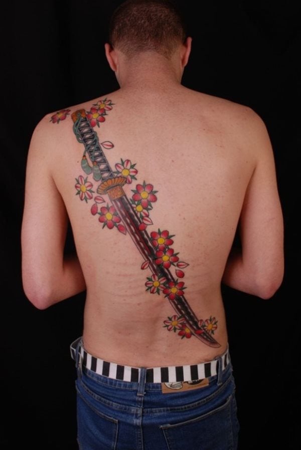 Tatuaje samurai de una espada enorme que cruza toda la espalda de manera diagonal y que se ha decorado con unas sencillas flores naranjas y amarillas, un bonito tatuaje que será de agrado para todos los amantes de este tipo de tattoos