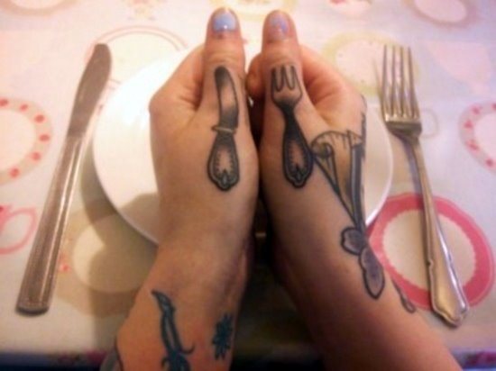 Divertido diseño de un cuchillo y tenedor sobre los pulgares, es un tatuaje simpático para los amantes de la mesa y para el que se ha utilizado como reflejo unos cubiertos señoriales de antaño y que han quedado genial en los pulgares de esta mujer
