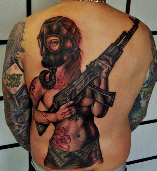 Tatuaje espectacular de una mujer desnuda que porta sobre sus manos una gran metralleta y a la que se le ha tatuado una máscara