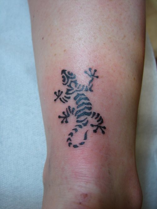 Disimulado tatuaje de un pequeño reptil que ha sido dibujado sobre dibujos tribales, uno de los tatuajes pequeños que más nos gustan, sobre todo por el relleno que se ha utilizado y las patas tan bien conseguidas