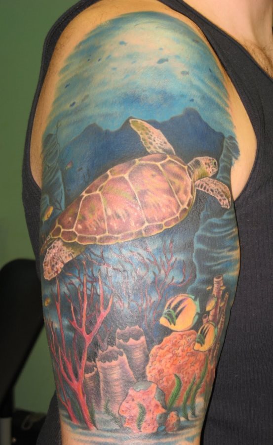 Espectacular diseño de gran tamaño que ocupa toda la parte superior del brazo, en el que se puede ver a una tortuga marina nadando en el fondo oceánico lleno de arrecifes, algas, esponjas y peces