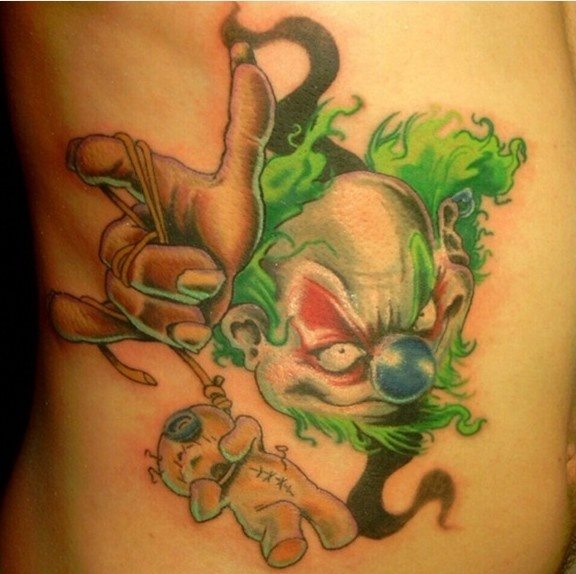 Payaso tétrico y de uñas largas que tiene sobre sus manos las cuerdas de un osito de peluche que le dan un toque fúnebre a este tatuaje del payas ocon el pelo verde y la nariz azul, un toque diferente el de la nariz azul ya que casi todos los payasos que acostumbramos a ver llevan la nariz roja