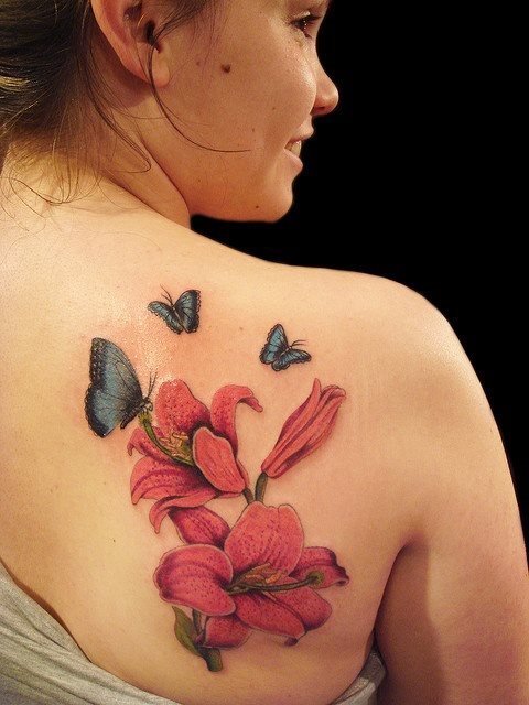 Realista y bonito tatuaje de flores y mariposas, en la que el complementar la gran mariposa que se posa en la flor con otras dos mariposas más pequeñas ha sido una gran idea porque le queda genial a esta chica el tatuaje sobre la espalda, gracias a la belleza y a la destreza del tatuador y por supuesto, al buen gusto de la tatuada
