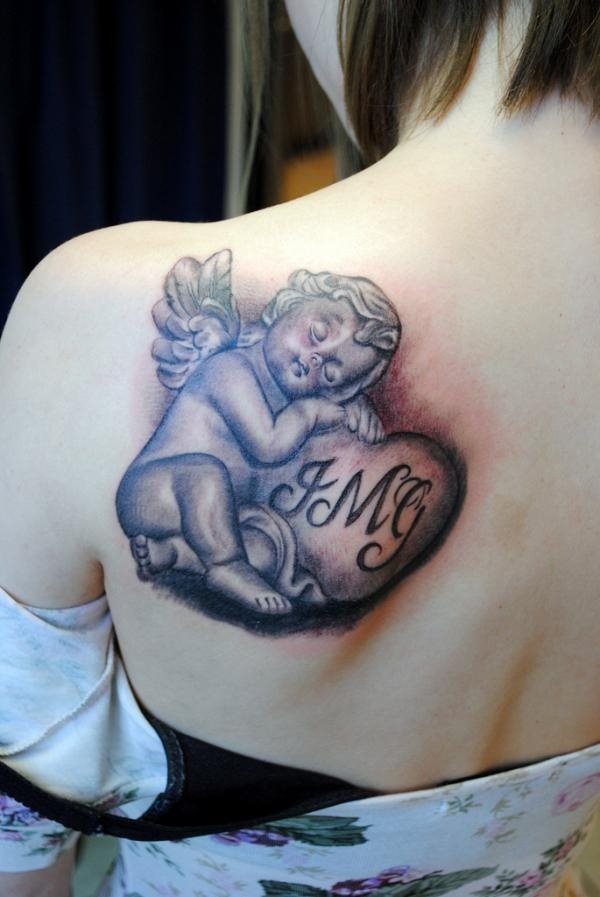 Tattoo en el hombro de un pequeño y regordete ángel con bonitos rizos que se encuentra dormido sobre un corazón al que se le han añadido las iniciales, posiblemente de los hijos o personas amadas por esta chica