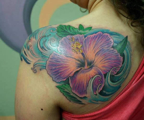 Tatuaje en el hombro y parte de la espalda de una flor de color lila con hojas verdes, cuyo acertado fondo está compuesto por un oleaje de color tuequesa que ha quedado fantásticamente bien en la espalda de esta mujer