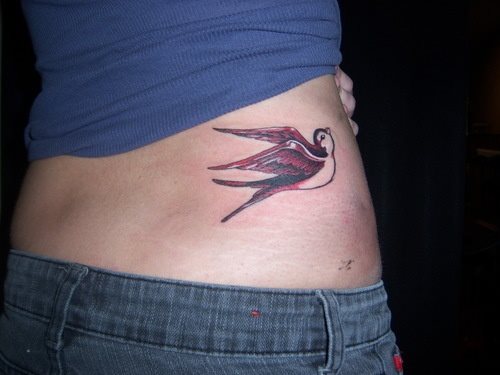 Tatuaje de una golondrina en negro y rojo sobre el costado de una mujer, los colores empleados son el rojo y el negro y la golondrina parece mirar hacia arriba como queriéndole decir algo a la mujer que lleva el tatuaje