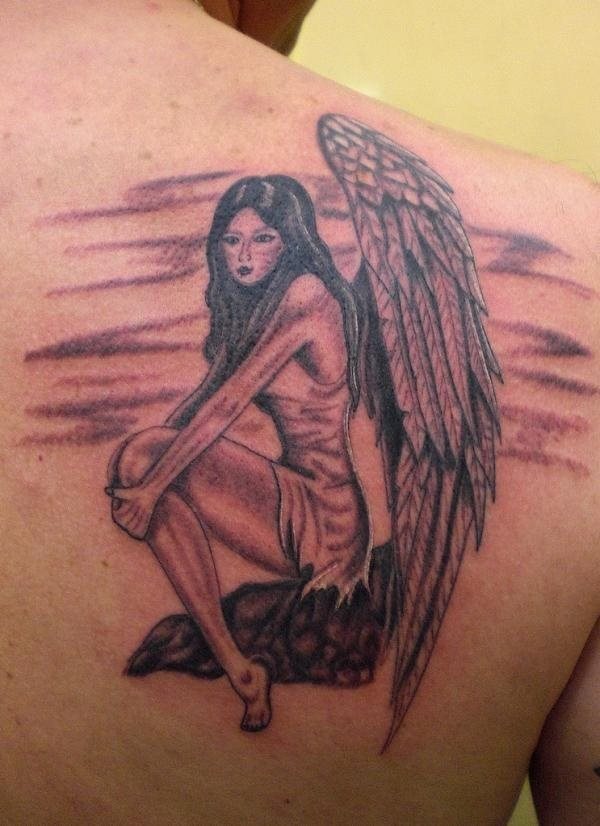 Tatauej de una mujer semi desnuda sentada sobe una piedra y a la que se le han tatuado unas grandes y bonitas alas con pluas muy largas, un tatuaje sencillo y que a la vez ha quedado muy bonito, al que nosotros tal vez no le hubiéramos dibujado los garabatos que hacen las veces de fondo